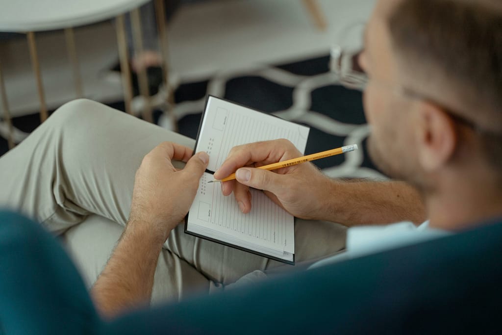 Servicios An adult man writing with a pencil in a notebook while seated indoors.